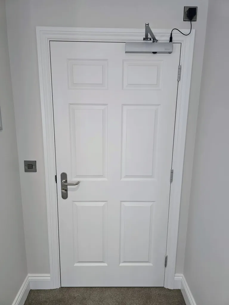 White single door with overhead door closer and metal handle in a plain wall setting