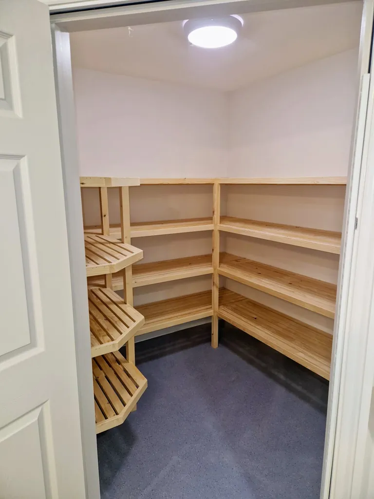 Small storage room with L-shaped wooden batten shelving and open door view